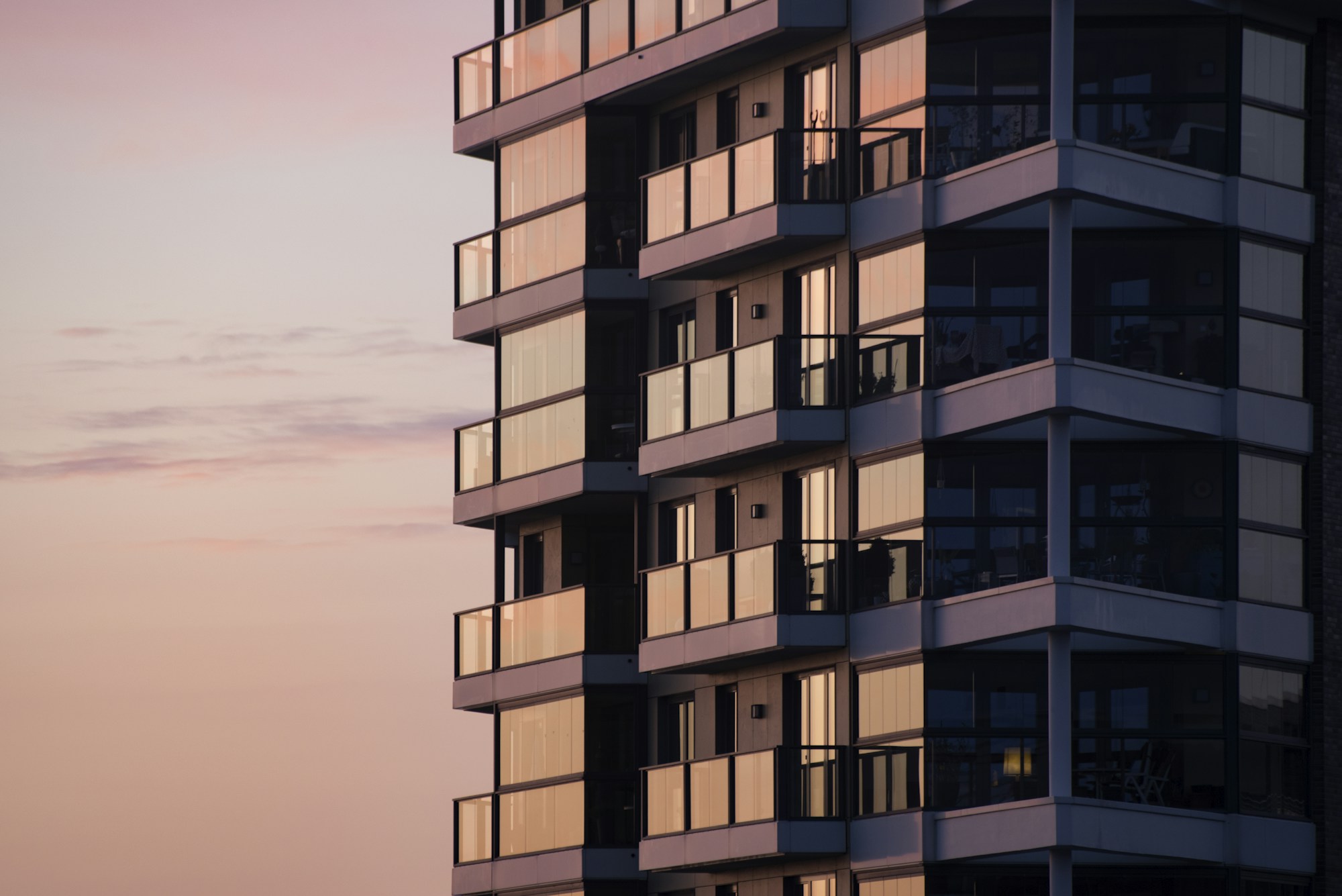 A modern apartment building with glass balconies reflecting a soft pink sky